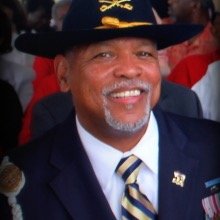 A smiling elderly man wearing a dark cowboy hat, a suit with a striped tie, and an American flag pin, attending a formal outdoor event.