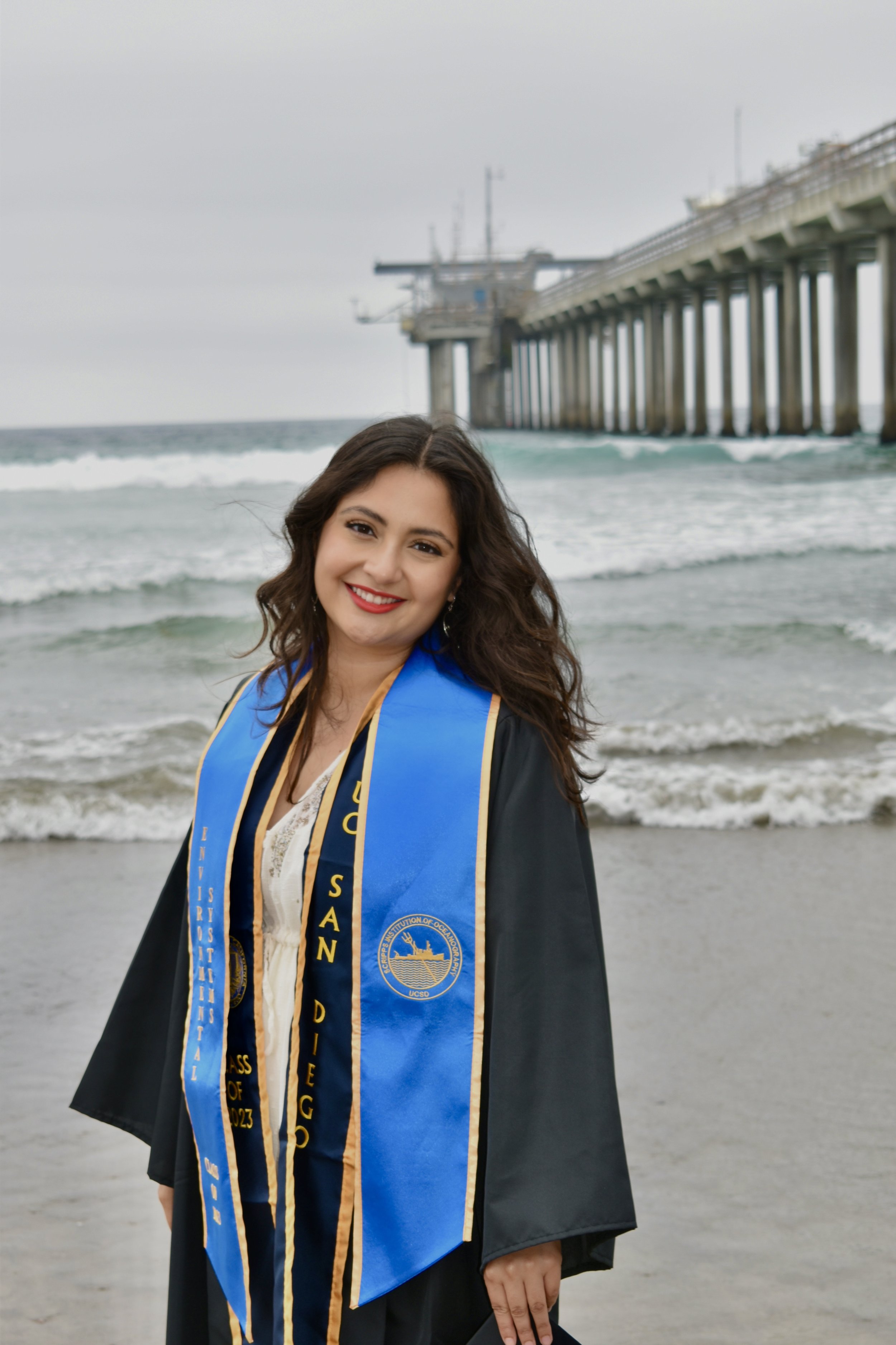 A young woman in graduation cap and gown with a blue and gold stole stands on a beach with the ocean and pier in the background.