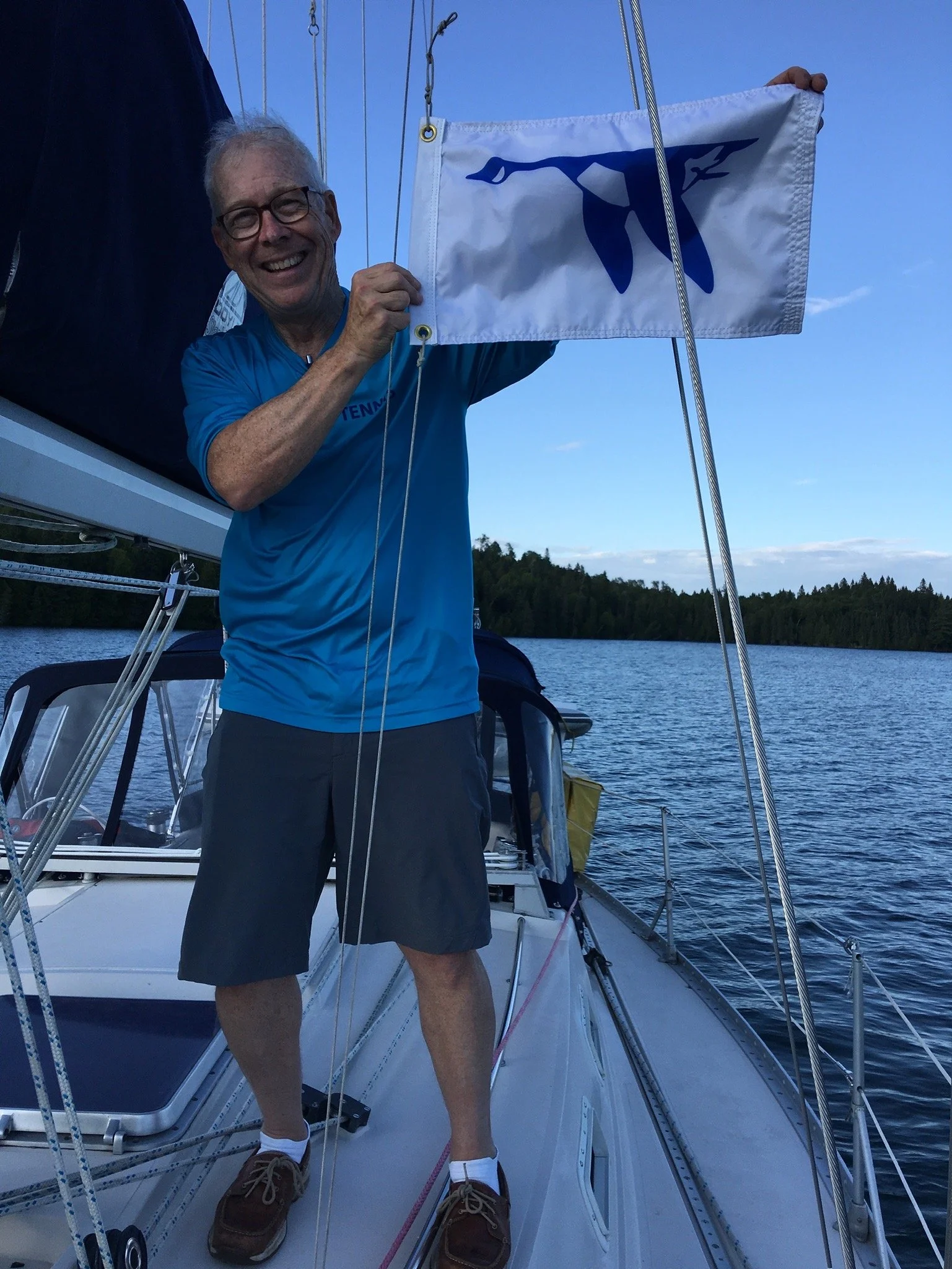 An elderly man smiling and holding a small flag on a boat, with water and trees in the background, during daylight.